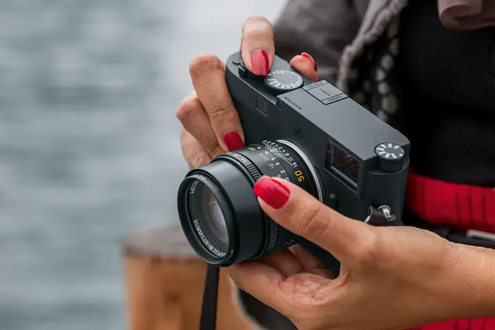 Leica M camera with 50 mm lens — Close-up of a person with red-painted nails holding a black Leica M rangefinder camera fitted with a 50 mm Summilux-M lens.