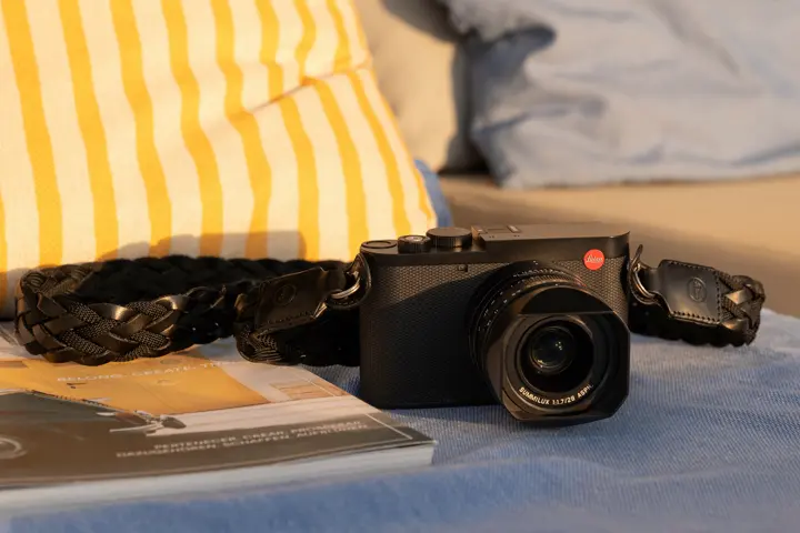 Leica Q camera with strap — Leica Q compact camera resting on a blue surface beside a black woven strap, photographed in warm natural light.