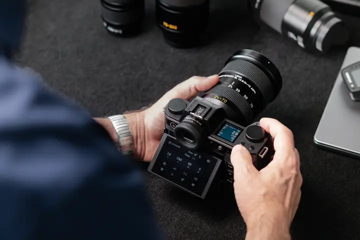 Person holding and operating a Leica SL-System camera fitted with a Leica Vario-Elmarit-SL 24–70 mm lens, with the rear display and top control screen visible on a dark tabletop.