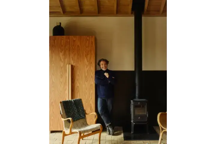 Jay Osgerby stands with his arms crossed beside a black wood-burning stove in a warmly lit interior with wooden walls and ceiling, with a woven armchair in the foreground.