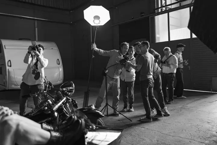 A black-and-white photograph of photographers experimenting with cameras and studio lighting around a motorcycle inside an industrial warehouse.