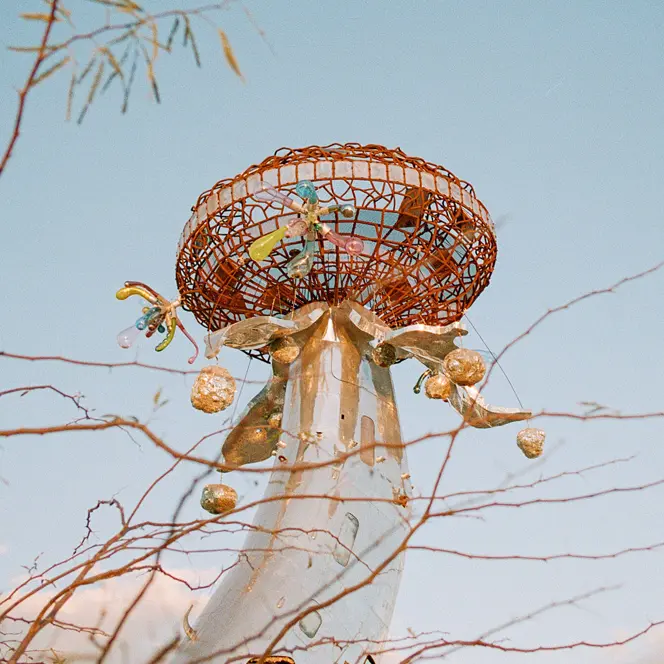 Metallic sculpture with colourful details in front of a blue sky and branches.