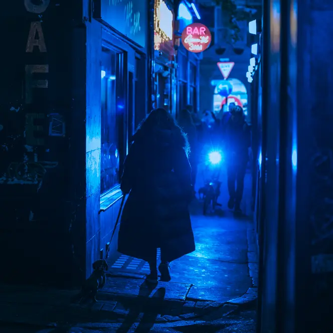 Person walking a dog through a narrow alley at night, illuminated by blue neon lights.