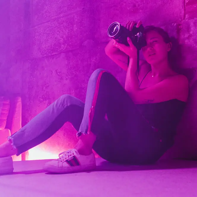 Alex Drewniak sits on the floor, taking pictures in a room lit by pink-purple light.