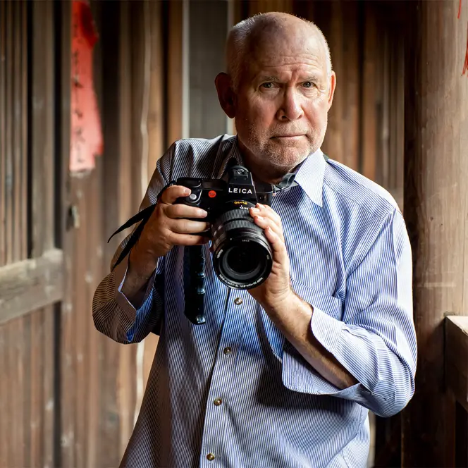 Steve McCurry holds a Leica camera and stands in front of a wooden house.