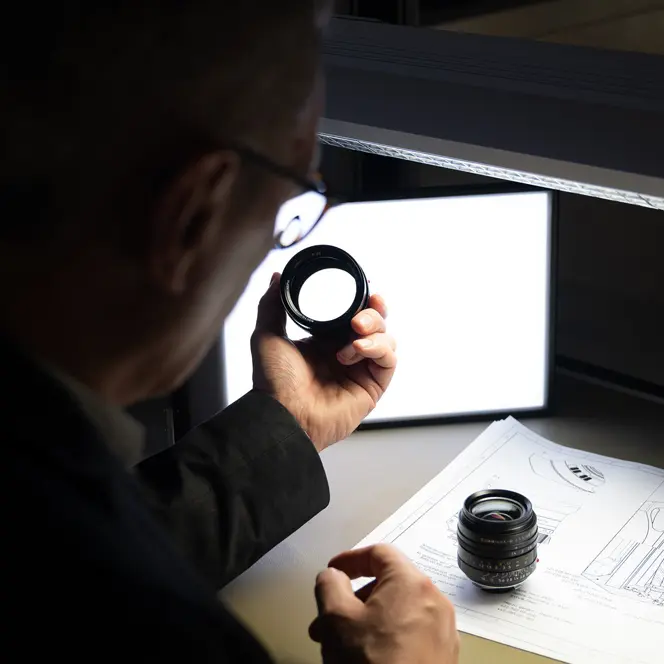 A man inspects a camera lens, technical drawings lie on the table.