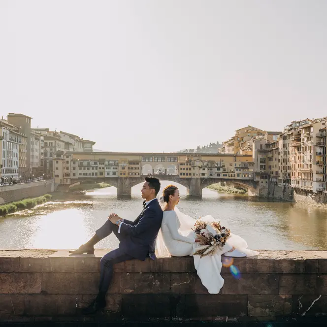 A wedding couple sits on a wall overlooking the river, surrounded by old buildings.