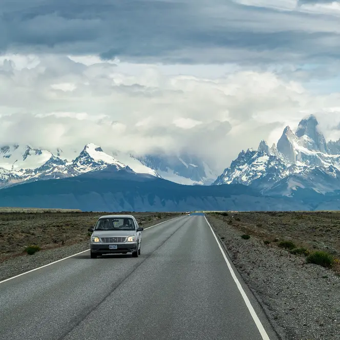 A car is driving on a road with snow-covered mountains in the background.