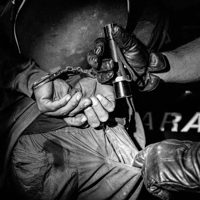 Close-up of a handcuffed person and another person searching his pants pocket with a flashlight.