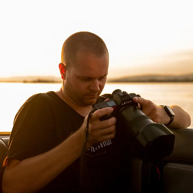 Tino Scherer looking at his camera. In the background is a sea at sunset.