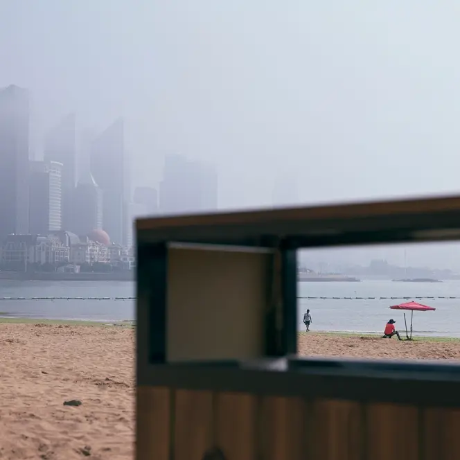 View through the rectangular opening of a garbage can on the beach with skyscrapers in the foggy background.