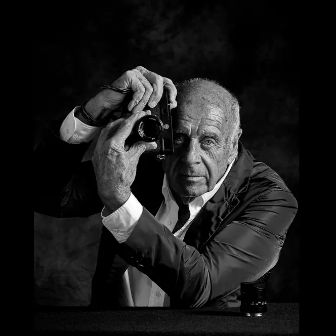 Elderly man holds a camera in front of his face and turns the lens of the camera.