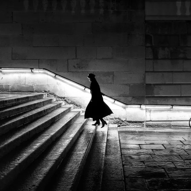 A woman in a long coat is walking up a staircase at night. The banister is illuminated.
