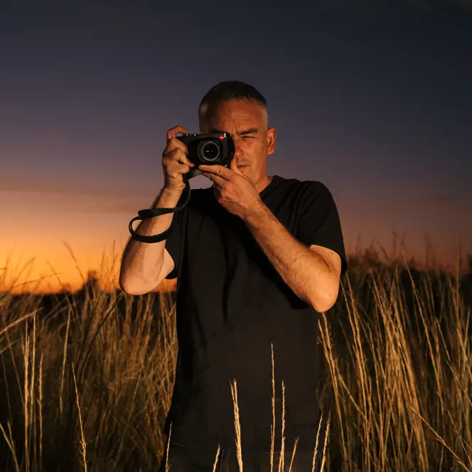 Person in a field with tall grass taking photos with a camera at sunset.