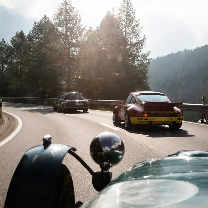 Vintage cars drive along a winding mountain road surrounded by trees and rocks.