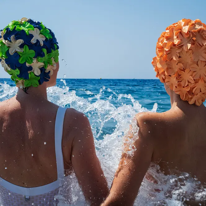 Two people in swimsuits and colorful bathing caps sitting in splashing water in the sea.