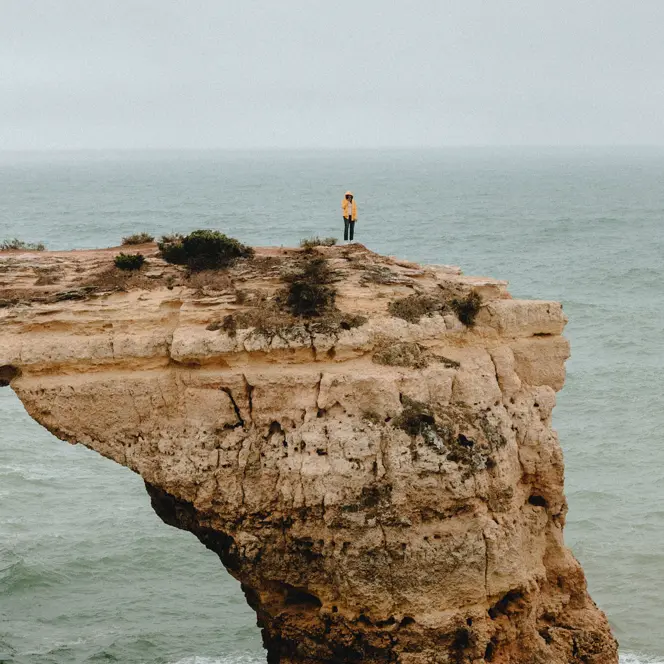 A person is standing alone on a rocky cliff jutting out into the ocean, and it is raining.