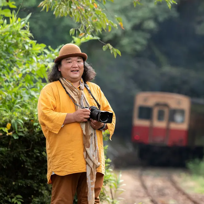 Man with camera stands beside railroad tracks and a train in background.