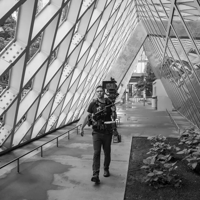 A cameraman walking with a professional camera setup in an architectural space.