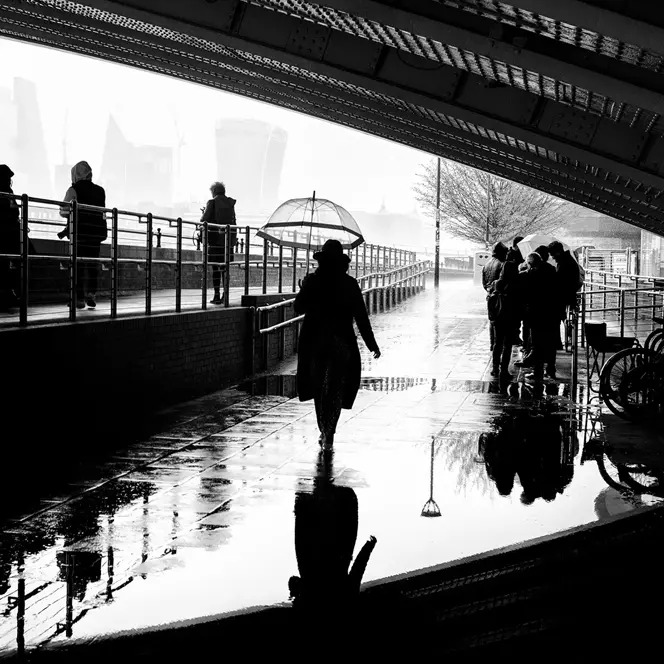 People walking under a bridge in the rain, silhouettes reflected in puddles.