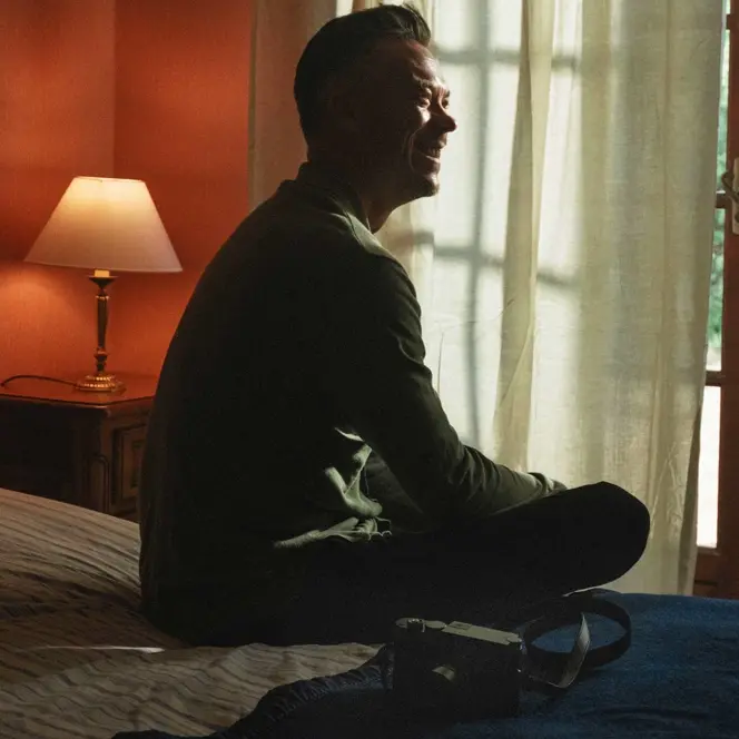 Smiling person sitting on a bed and a camera lying next to it.