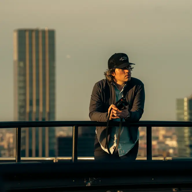 A man holding a camera leans against a railing with skyscrapers in the background.