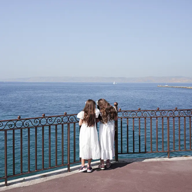 Two girls in white dresses stand at a railing and look to the sea.