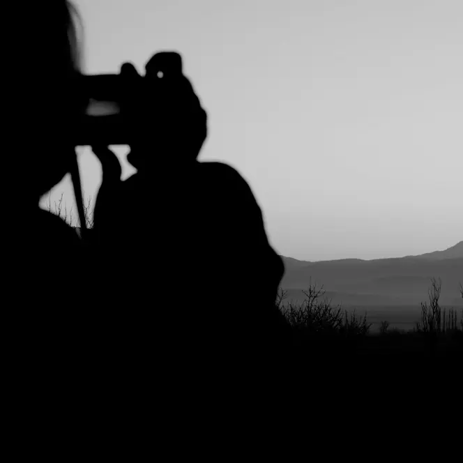 Black-and-white photograph of a person taking a picture of a lake and mountains in the background.