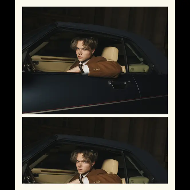 A person in a brown jacket sits in a car and looks through the open window.