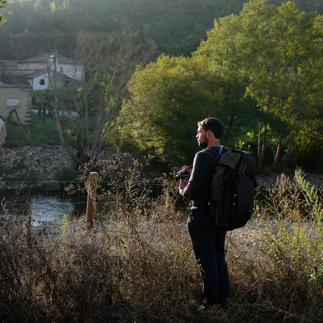 A man with a rucksack stands in the countryside and looks out over a small settlement by the river