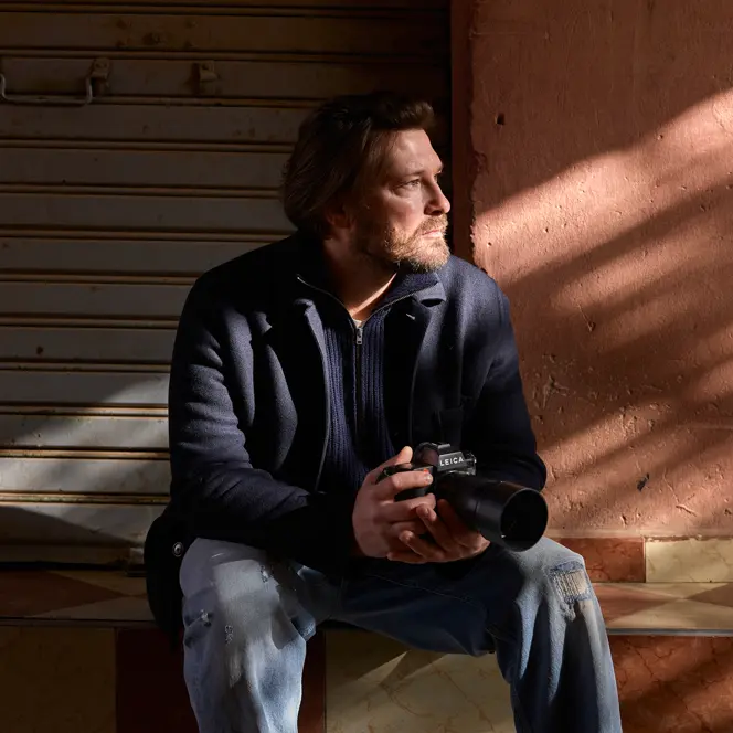 A man sits on a step with a camera in his hands, his face in the sunlight, in front of a closed shutter.