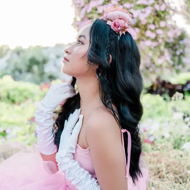 Woman in a pink dress and white gloves is sitting in a rose garden looking to the side.