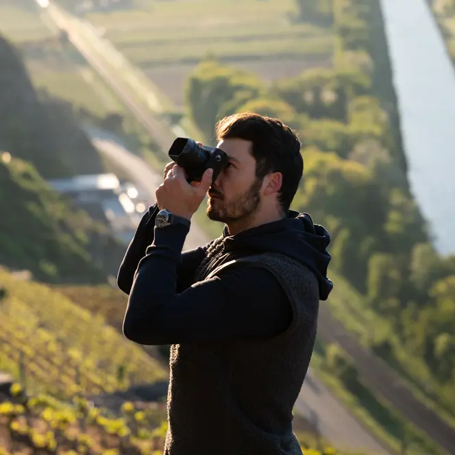 A photographer takes a picture on a sunlit vineyard with a river and trees in the background.