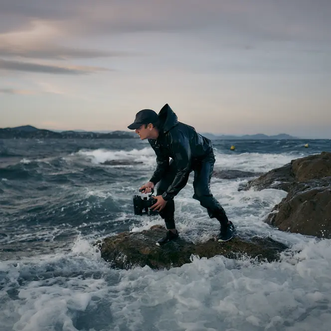 A person is standing on a rock in a stormy sea, holding a pair of binoculars.