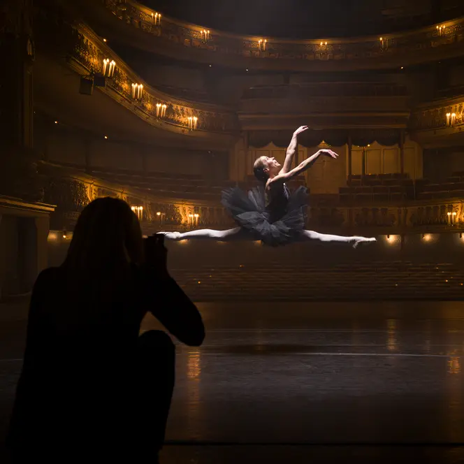 A person in the shade takes a picture of a ballerina jumping in an illuminated theatre.