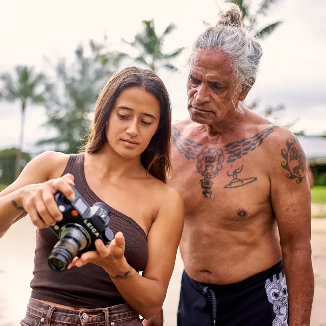 Mainei Kinimaka shows pictures on a camera to a man with Hawaiian tattoos on his upper body