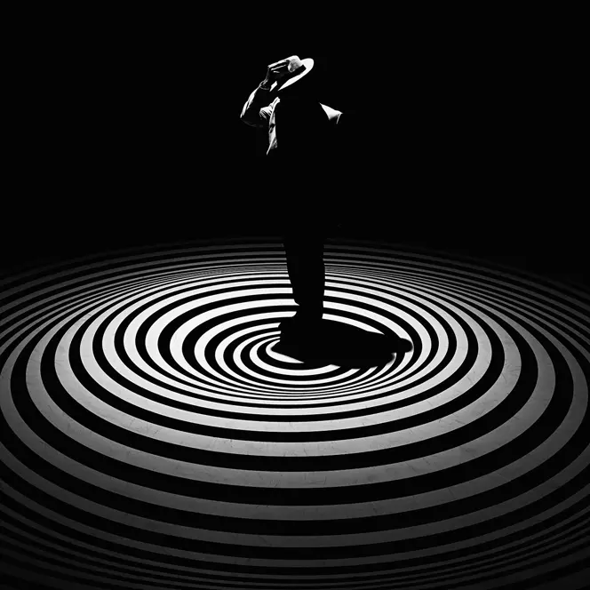 A single person with a hat stands in the centre of concentric circles on a dark background