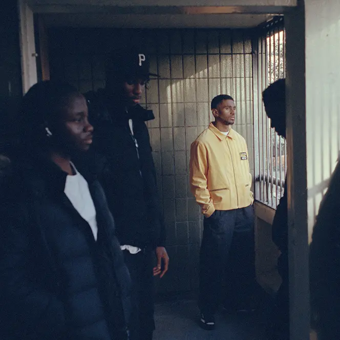 Five men in a shady hallway looking outside, the one in the middle wearing a yellow jacket.