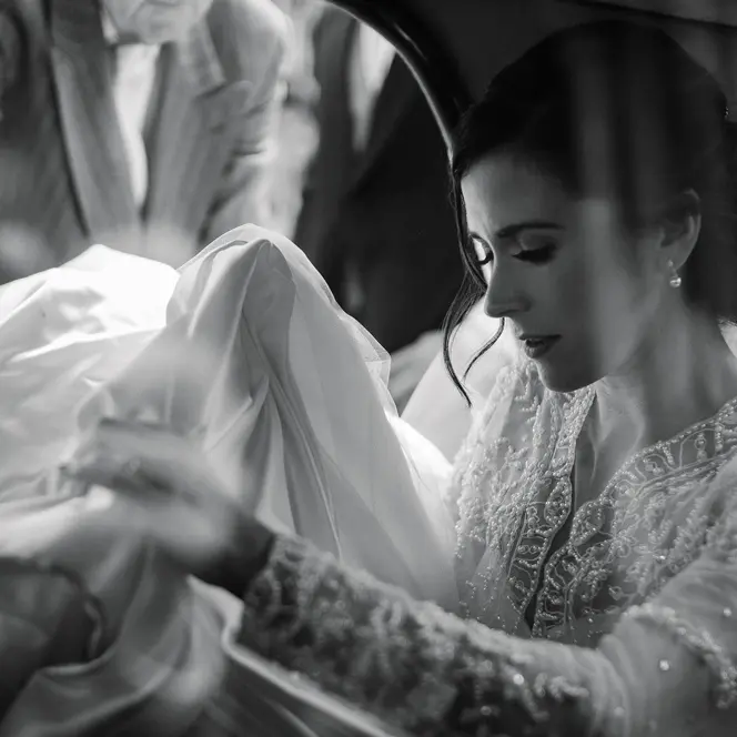 A bride sitting in a car holding her veil and a man standing next to the open car door.