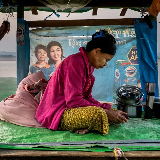 Woman sits on plastic tarp looking at a phone, in background are other people and a river.