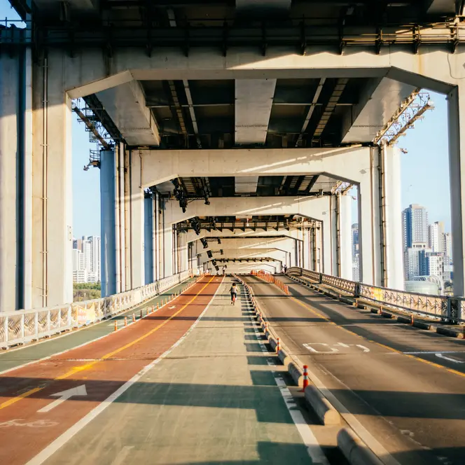 Single cyclist on a bridge with a separate cycle and pedestrian path and road, framed by tall city buildings in the background.