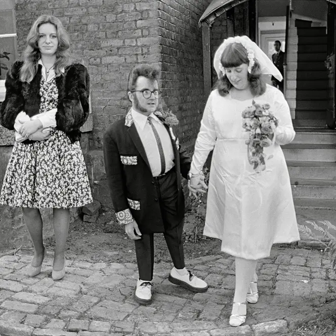 A wedding couple and a woman in a dress stand in front of a building.
