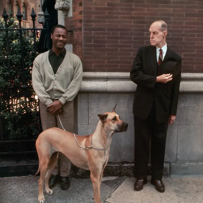 Two men, one with a dog on a lead, stand in front of a brick wall.
