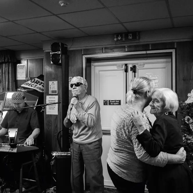 A man sings into a microphone while two older women dance and a man plays the keyboard in the background.