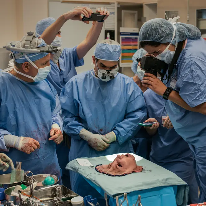 A group of people in surgical gowns look at a mask representing a face, and some take pictures of it.