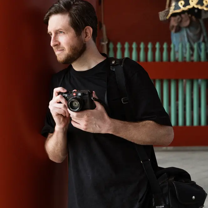 A photographer holds a camera while standing near traditional architecture, with two people in kimonos in the background.