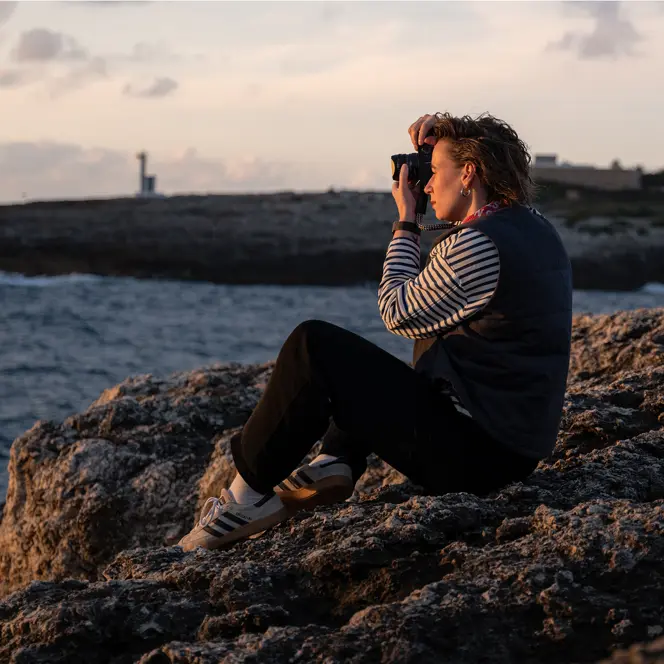 Julia Nimke is sitting on a rock by the sea with her Leica camera, taking photos