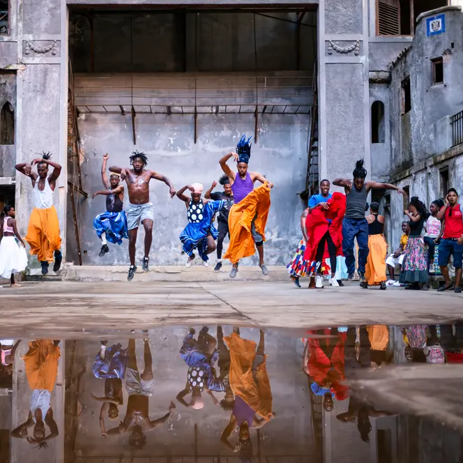 Traditional dancers performing in a historic courtyard in Cuba, captured mid-jump with vibrant costumes and reflections in a rain puddle.
