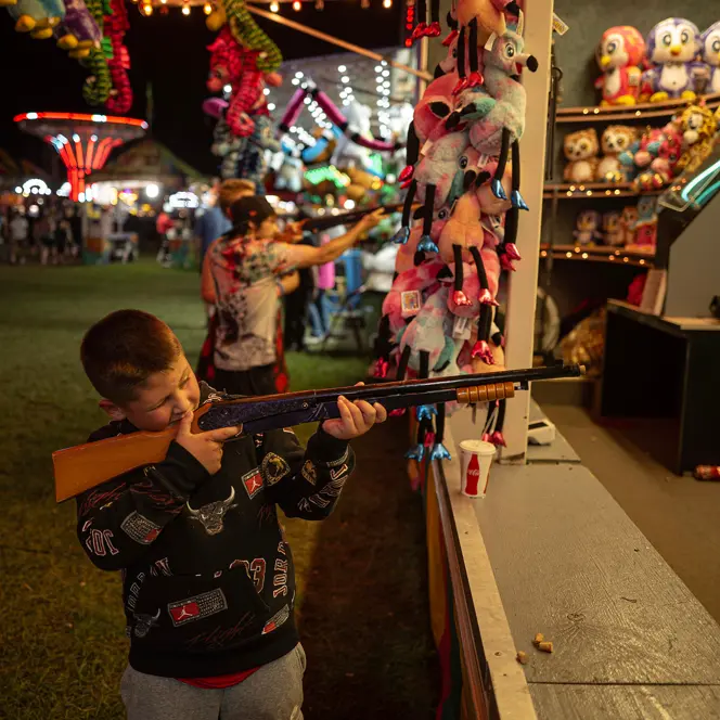 Child aiming at a carnival shooting game, capturing the vibrant atmosphere of a nighttime fairground and family entertainment.