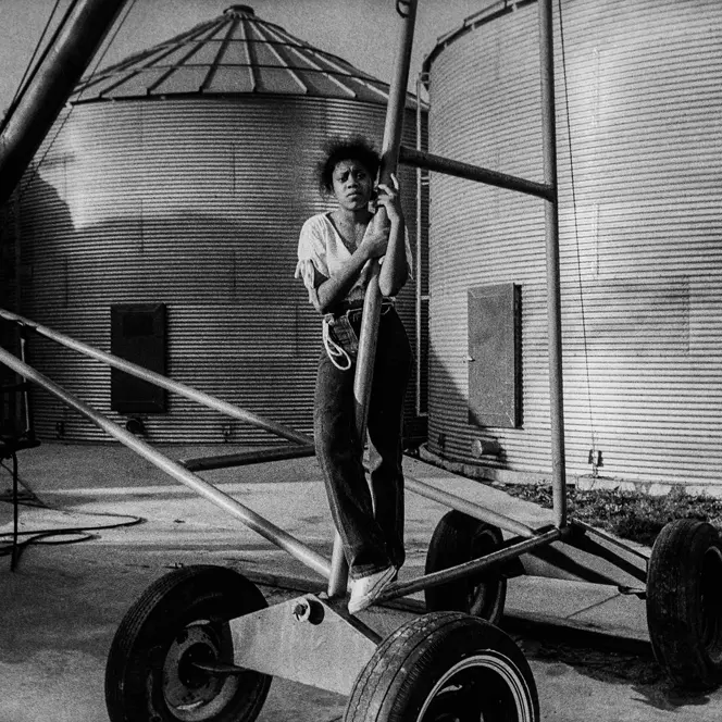 Young girl standing beside farm equipment and grain silos on a family farm in Arkansas, captured in black and white. A documentary photograph reflecting rural life, agricultural heritage, and generational connection.